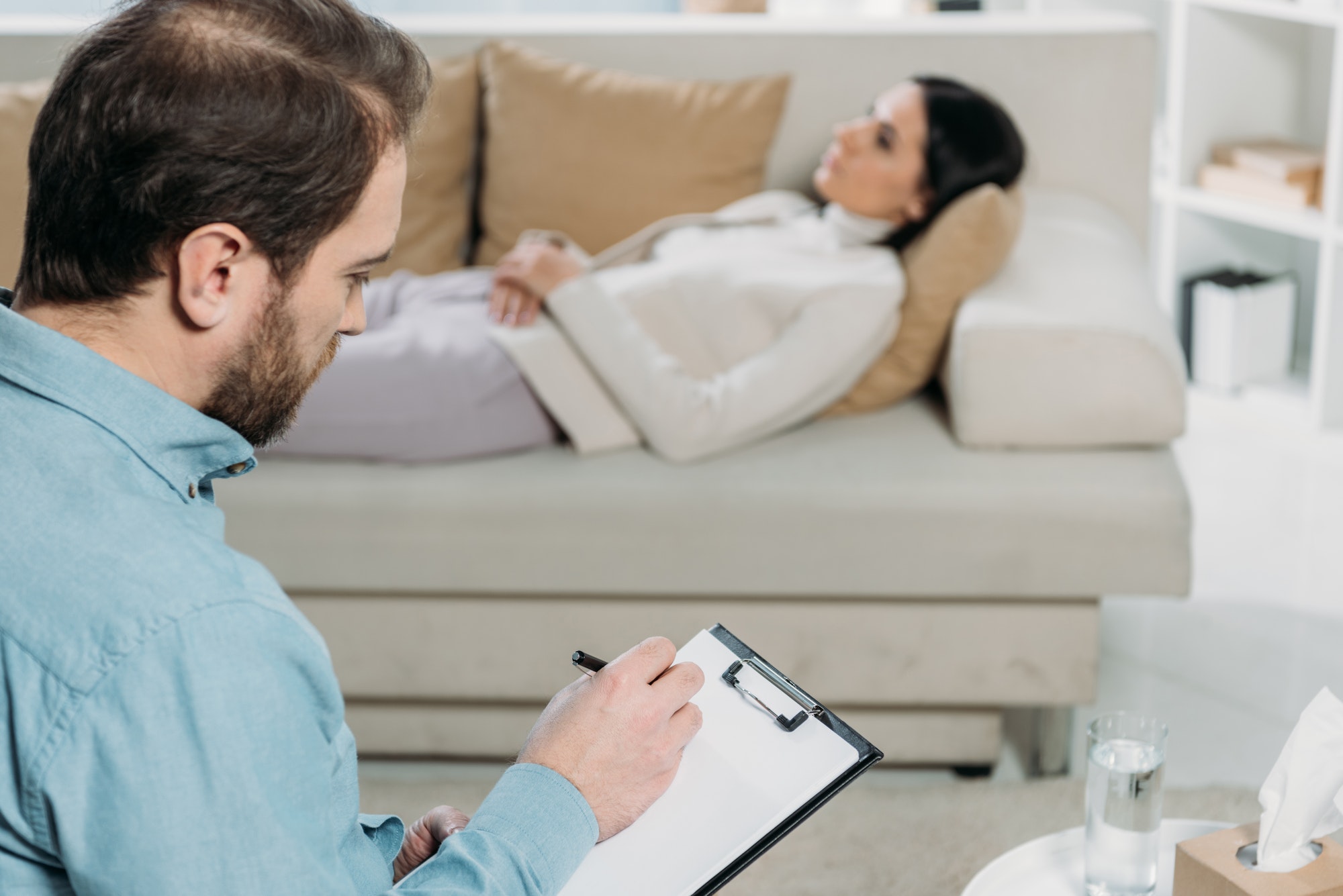 bearded psychotherapist writing on clipboard while upset patient lying on couch
